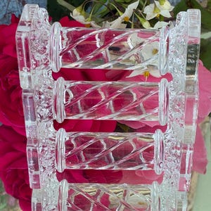 May include: Clear glass decorative object with a repeating cylindrical design. The cylinders have a spiral pattern and are connected by decorative, rectangular supports. The background features pink roses and white flowers.