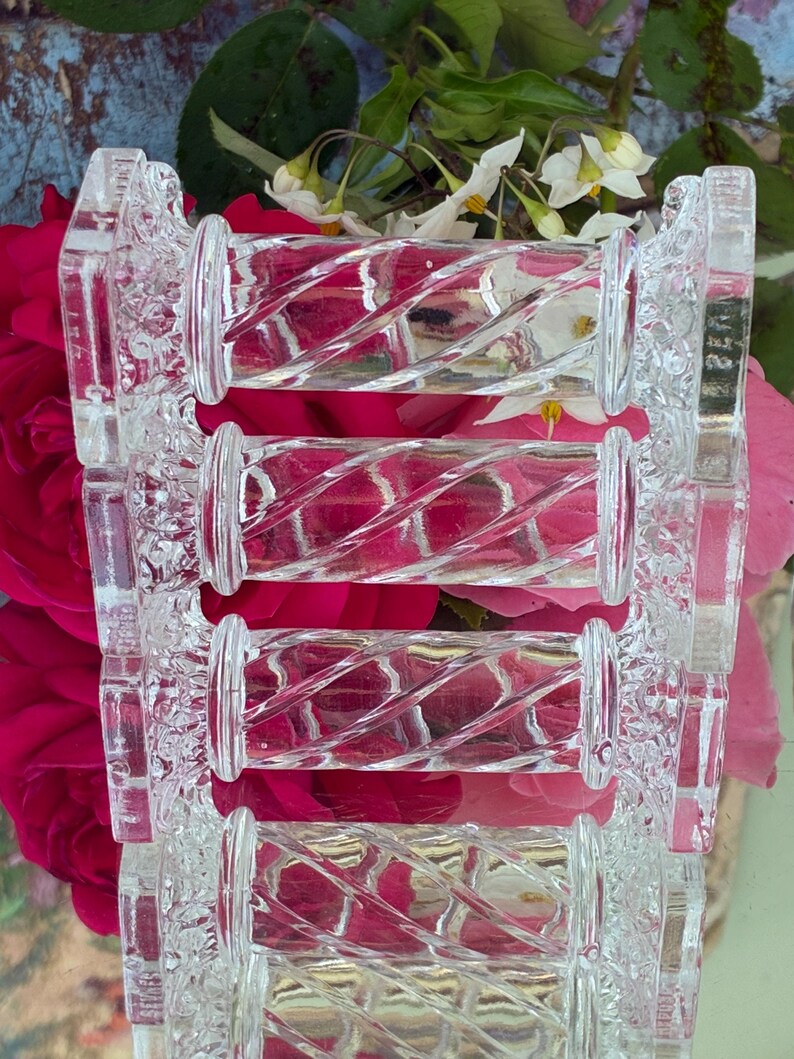 May include: Clear glass decorative object with a repeating cylindrical design. The cylinders have a spiral pattern and are connected by decorative, rectangular supports. The background features pink roses and white flowers.