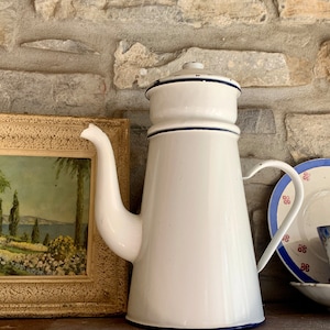Könnte beinhalten: Weiße Email-Kaffeekanne mit blauem Rand und Ausguss. Die Kanne steht auf einem Regal mit einem gerahmten Bild und einem kleinen Teller und einer Tasse.