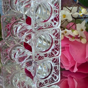 May include: A stack of clear, square glass napkin rings with a decorative, swirling design. The rings have a square frame with an ornate pattern. The background includes pink roses and white flowers.