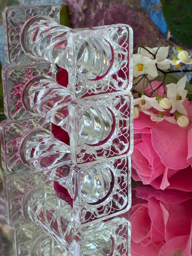 May include: A stack of clear, square glass napkin rings with a decorative, swirling design. The rings have a square frame with an ornate pattern. The background includes pink roses and white flowers.