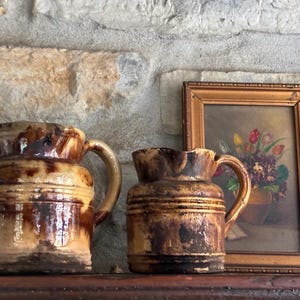 May include: Two antique stoneware pitchers with handles, featuring a brown and tan glaze. The pitchers are displayed on a wooden shelf, with a framed painting of flowers in the background. The pitchers have a rustic, vintage aesthetic.