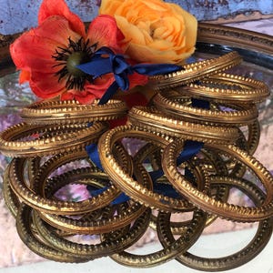 May include: A collection of antique gold-colored napkin rings with a textured design. The rings are arranged in a pile, some tied with a dark blue ribbon. A vibrant orange and red flower arrangement sits in the background.
