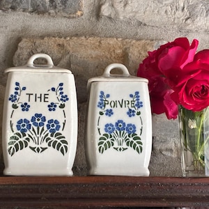 Op de afbeelding: Twee crèmekleurige keramische potten met deksels, met de tekst "THE" en "POIVRE" in blauwe letters, versierd met blauwe bloemmotieven. Rechts staat een vaas met rode rozen. De potten staan op een donkere houten plank.