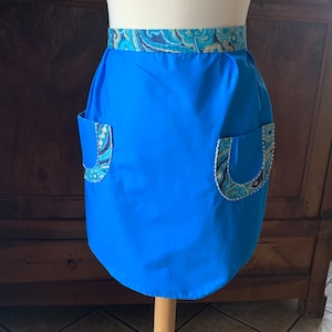 Pode incluir: Um avental azul com uma cintura com padrão paisley e dois bolsos. Os bolsos têm uma borda branca e um forro com padrão paisley.