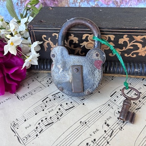 May include: An antique, weathered padlock with a key attached by a green string rests on sheet music. The lock is gray with red accents and a dark brown handle. A dark box with gold floral designs and a vibrant pink flower are in the background.