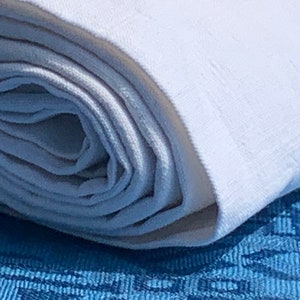 May include: A close-up shot of a rolled white fabric, possibly linen or cotton, showing its texture. The fabric rests on a blue patterned surface, creating a contrast. The rolled fabric suggests a textile product, such as a tablecloth or bedding.