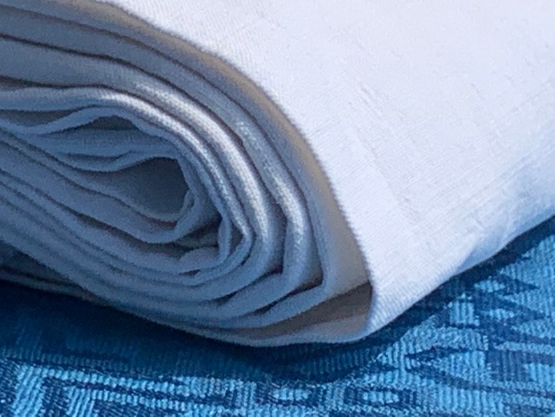 May include: A close-up shot of a rolled white fabric, possibly linen or cotton, showing its texture. The fabric rests on a blue patterned surface, creating a contrast. The rolled fabric suggests a textile product, such as a tablecloth or bedding.