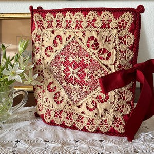 Può includere: Un cuscino decorativo rosso e crema con intricati dettagli in pizzo. Il cuscino presenta un design quadrato con un motivo a diamante centrale e un nastro rosso abbinato legato a un fiocco. Il cuscino è posto su una tovaglia di pizzo bianco.