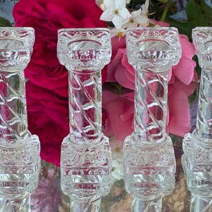 May include: Clear glass candle holders with a twisted column design. The candle holders have a square top and base with decorative detailing. The background features pink and white flowers.