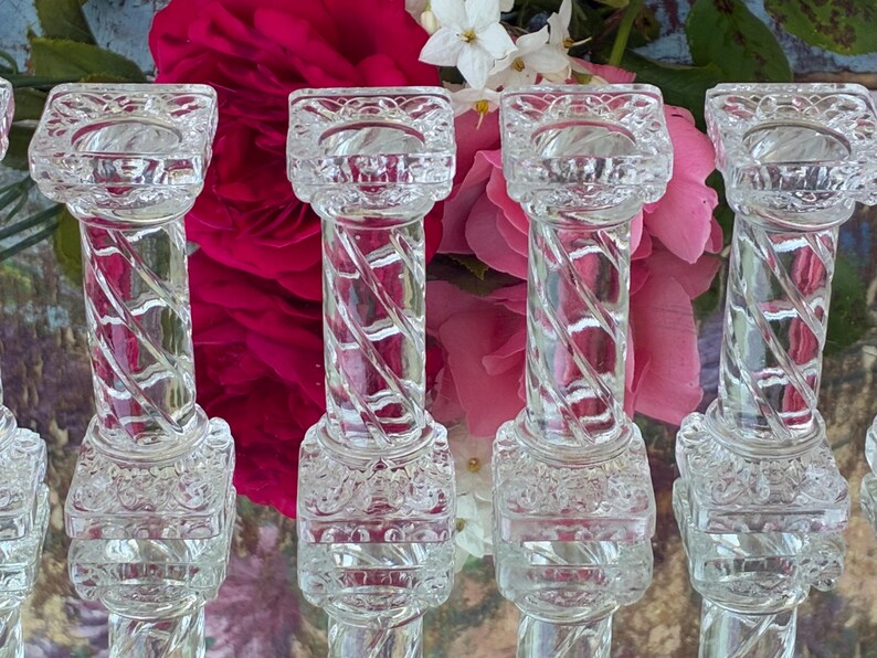 May include: Clear glass candle holders with a twisted column design. The candle holders have a square top and base with decorative detailing. The background features pink and white flowers.