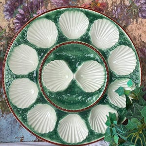 May include: A round, green and white ceramic oyster serving platter. The platter features a central well and surrounding shell-shaped compartments. The rim is green with a woven texture, and the edges are trimmed in red.