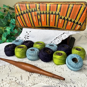 Peut inclure: Une boîte métallique ancienne avec un motif géométrique orange, jaune et noir est placée au-dessus d'une collection de bobines de fil colorées. Un rouleau à pâtisserie en bois repose sur une nappe en dentelle blanche.