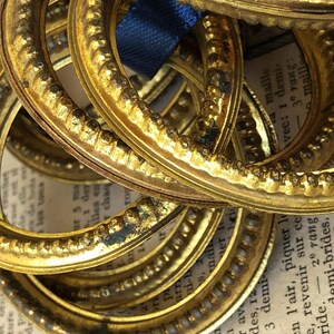 May include: A collection of antique, gold-colored oval frames with ornate detailing. The frames are stacked on top of each other, with a blue ribbon visible. The background includes aged paper with French text.