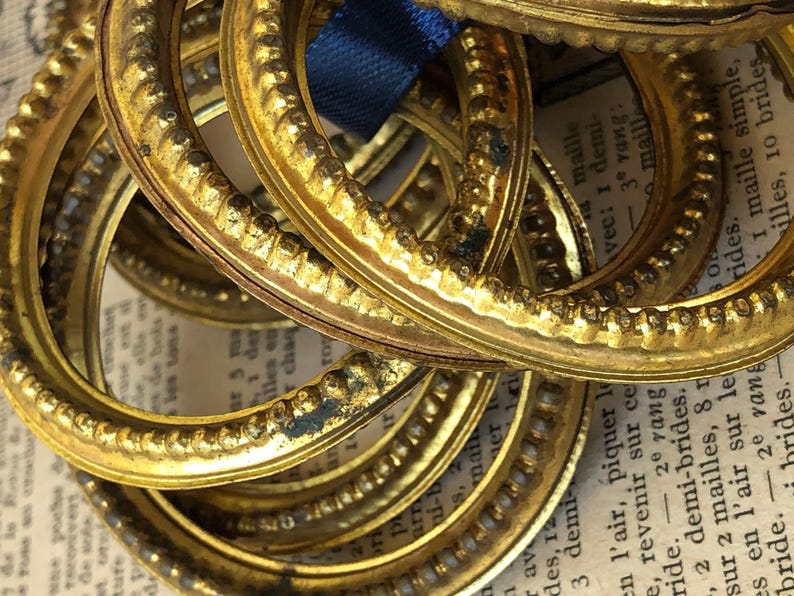 May include: A collection of antique, gold-colored oval frames with ornate detailing. The frames are stacked on top of each other, with a blue ribbon visible. The background includes aged paper with French text.