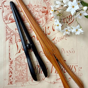 Può includere: Due fermapagina musicali per pianoforte antichi, uno in legno chiaro e l'altro in metallo nero. Sono appoggiati su una pagina di spartito vintage con testo e illustrazioni e fiori bianchi. Il legno misura circa 30 cm di lunghezza.