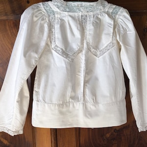 Può includere: Blusa bianca in cotone con maniche lunghe e bordino in pizzo intorno al collo e ai polsini.
