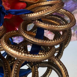 May include: A collection of ornate, gold-colored metal rings with a textured, beaded design. A dark blue ribbon weaves through the rings, and colorful flowers are visible in the background. The rings have a vintage or antique aesthetic.