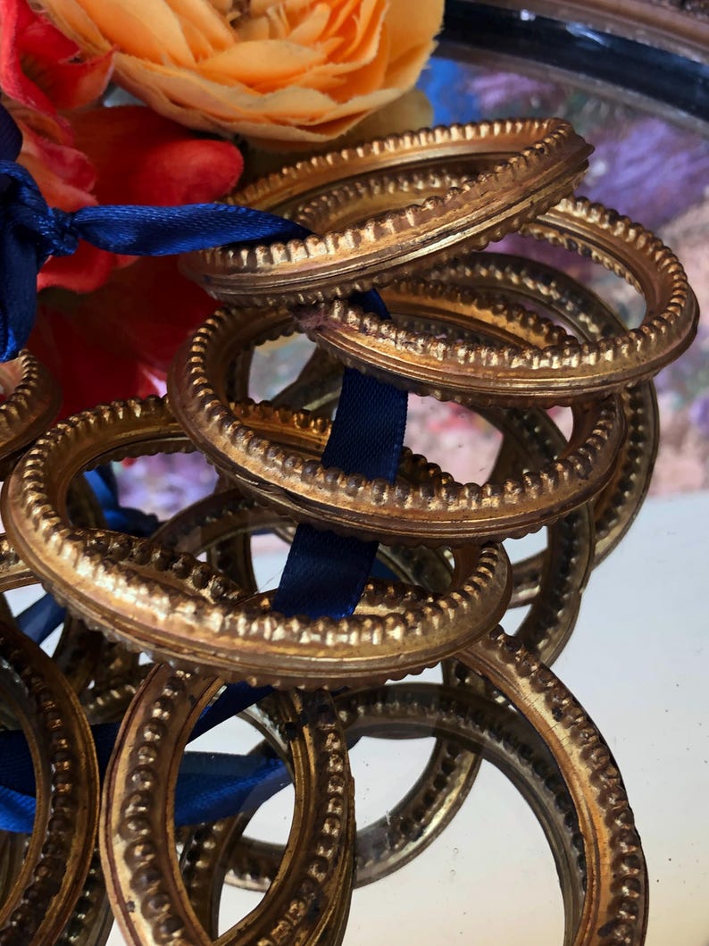 May include: A collection of ornate, gold-colored metal rings with a textured, beaded design. A dark blue ribbon weaves through the rings, and colorful flowers are visible in the background. The rings have a vintage or antique aesthetic.
