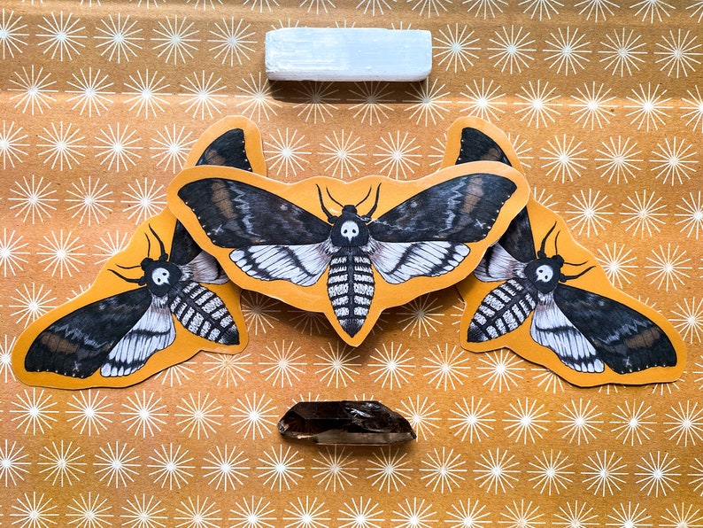 May include: Three black and white moth stickers on a brown background with a white starburst pattern. The moths have a skull and crossbones design on their wings.