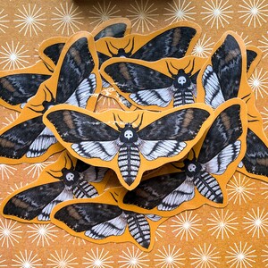 May include: A collection of black and white moth stickers with a skull design on a yellow background with white starburst patterns.