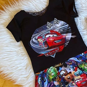 Könnte beinhalten: Schwarz-rotes Kurzarm-Shirt mit einem Cars-Druck, der Lightning McQueen auf einer Rennstrecke zeigt. Das Shirt wird mit einem schwarz-bunten Rock mit einem Cars-Druck mit verschiedenen Figuren kombiniert.