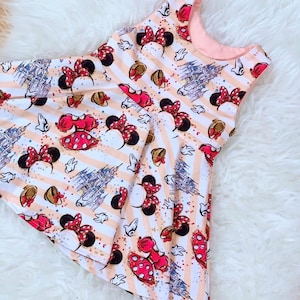 Könnte beinhalten: Ärmelloses Kleid mit einem Muster aus Minnie Mouse-Schleifen, gepunkteten Kleidern und Schlossillustrationen auf einem gestreiften Pfirsich- und weißen Hintergrund. Das Kleid hat ein rosa Futter und einen ausgestellten Rock.