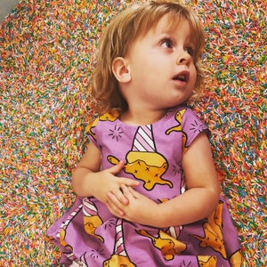 Könnte beinhalten: Ein junges Mädchen trägt ein lila Kleid mit einem gelben Eiswaffendesign. Das Kleid hat einen weißen Kragen und kurze Ärmel. Das Mädchen sitzt auf einer bunten Oberfläche.