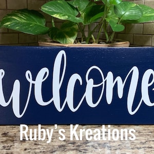 May include: A navy blue wooden sign with the word "welcome" written in white cursive lettering. The sign is made by Ruby's Kreations.