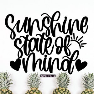以下が含まれることがあります： 「Sunshine state of mind」という黒と白のグラフィックテキストで、黒いハートが2つと太陽のグラフィックが描かれています。テキストは、下部に緑色のパイナップルが3つある白い背景に表示されています。