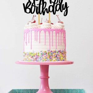 Puede incluir: Un pastel rosa con glaseado blanco y glaseado rosa derretido. El pastel est&aacute; decorado con chispas de colores y cubierto con una decoraci&oacute;n de pastel negra de "Feliz cumplea&ntilde;os" y cerezas rojas.