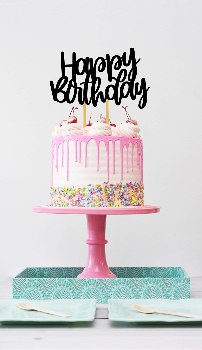 Puede incluir: Un pastel rosa con glaseado blanco y glaseado rosa derretido. El pastel est&aacute; decorado con chispas de colores y cubierto con una decoraci&oacute;n de pastel negra de "Feliz cumplea&ntilde;os" y cerezas rojas.