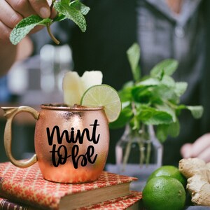 Puede incluir: Taza de cobre con una inscripci&oacute;n negra que dice "mint to be". La taza est&aacute; llena de un l&iacute;quido marr&oacute;n y adornada con una rodaja de lima y menta fresca. La taza est&aacute; sobre una pila de libros con una lima verde y un trozo de jengibre en el fondo.