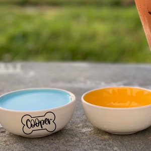 K&ouml;nnte beinhalten: Zwei Keramik-Haustier-Schalen, eine blaue und eine gelbe, mit wei&szlig;em Boden. Die blaue Schale hat den Namen "Cooper" in Schwarz darauf geschrieben.