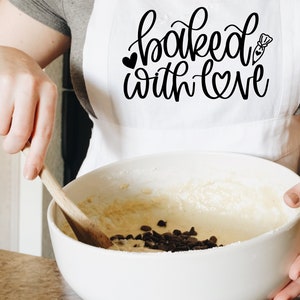 May include: A white apron with the text "baked with love" in black script. A person is wearing the apron and stirring a bowl of batter with chocolate chips.