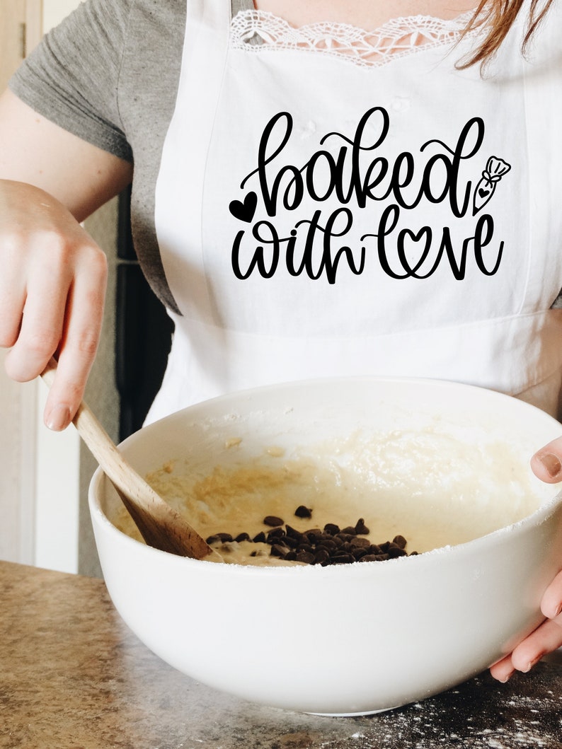 May include: A white apron with the text "baked with love" in black script. A person is wearing the apron and stirring a bowl of batter with chocolate chips.