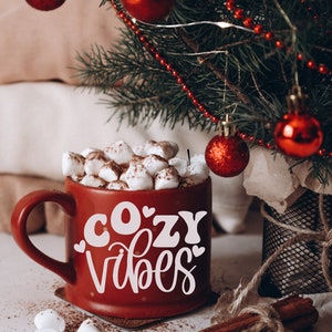 Puede incluir: Una taza roja con letras blancas que dicen "cozy vibes" llena de chocolate caliente y malvaviscos. La taza est&aacute; sobre una superficie blanca con palitos de canela y un peque&ntilde;o &aacute;rbol de Navidad en el fondo.