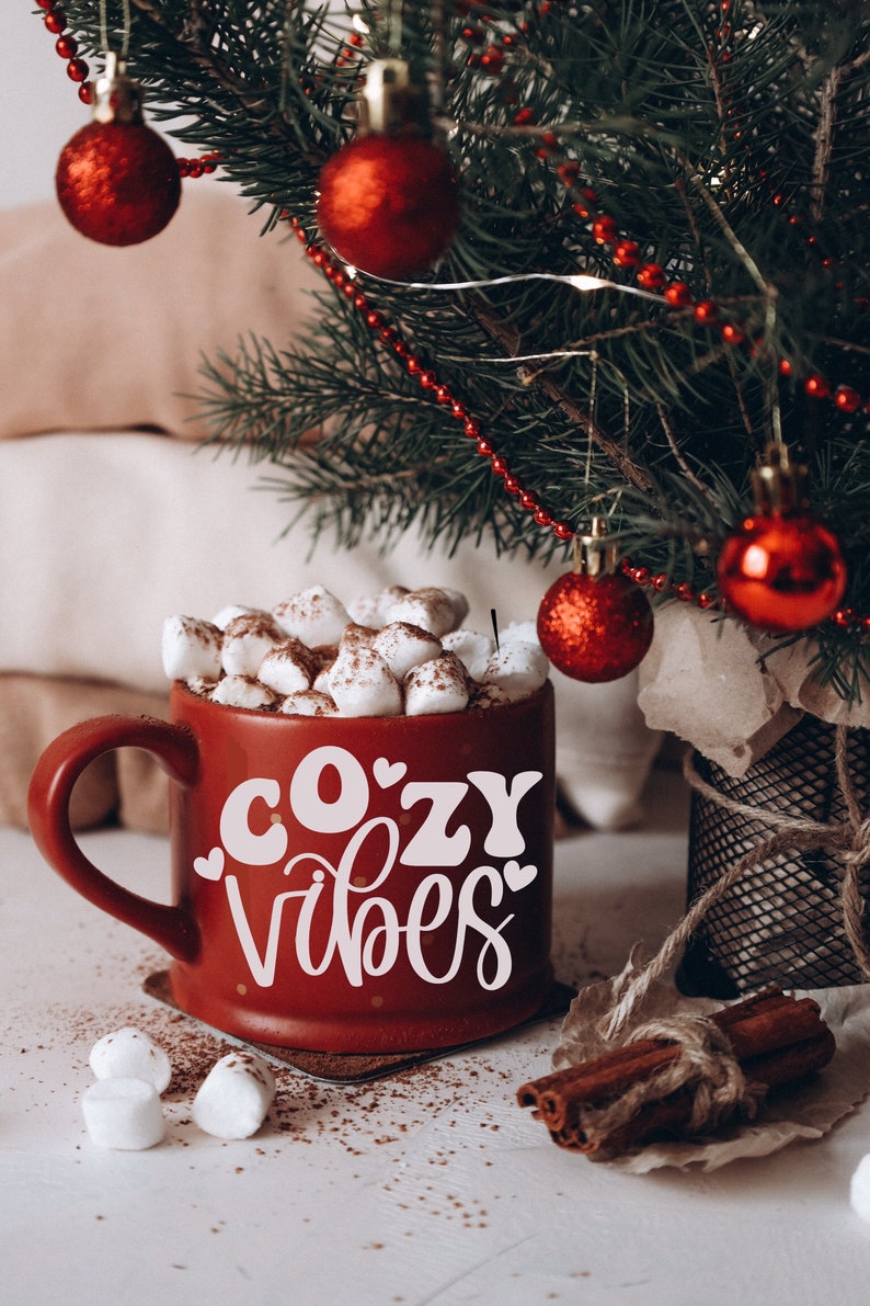 Puede incluir: Una taza roja con letras blancas que dicen "cozy vibes" llena de chocolate caliente y malvaviscos. La taza est&aacute; sobre una superficie blanca con palitos de canela y un peque&ntilde;o &aacute;rbol de Navidad en el fondo.