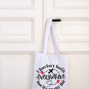 May include: White canvas tote bag with black text that reads "I haven't been everywhere but it's on my list". The text is in a circle with a plane and red location pins.