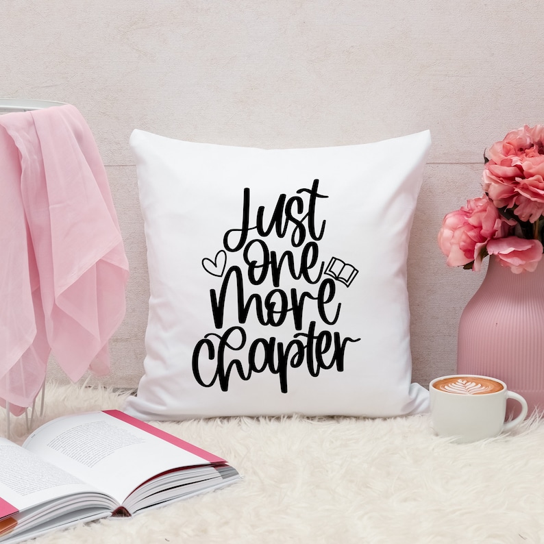 May include: White throw pillow with black text that reads "Just one more chapter". The pillow is on a white fluffy surface with a book and a cup of coffee.