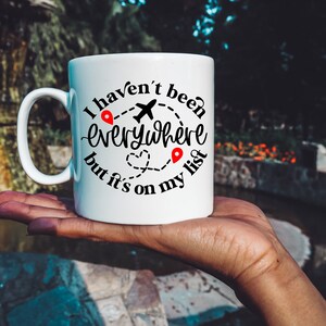 May include: White ceramic mug with a black graphic that reads "I haven't been everywhere but it's on my list". The graphic includes a plane, a heart, and two red location pins.