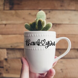 May include: White ceramic mug with a black cursive inscription that reads "Thank You" with a heart symbol. The mug is filled with a small green cactus plant.