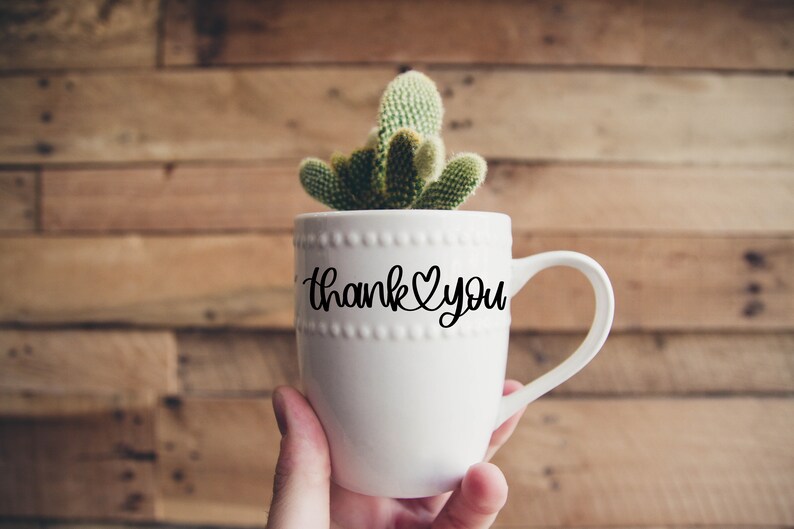May include: White ceramic mug with a black cursive inscription that reads "Thank You" with a heart symbol. The mug is filled with a small green cactus plant.
