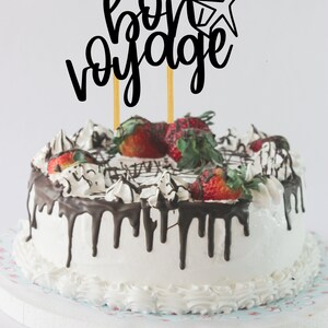 May include: A white cake with chocolate frosting and strawberries, topped with a black cake topper that says "bon voyage" with a small airplane graphic.