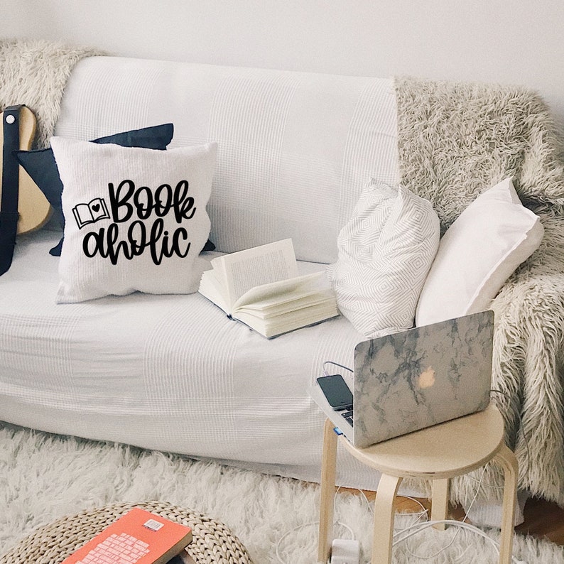 May include: White couch with a throw pillow that says "Bookaholic" in black lettering. A book, a laptop, and a small side table are on the couch.