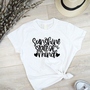 以下が含まれることがあります： 「Sunshine state of mind」という黒い文字が書かれた白いTシャツ。小さな太陽のグラフィックと2つのハートが描かれている。