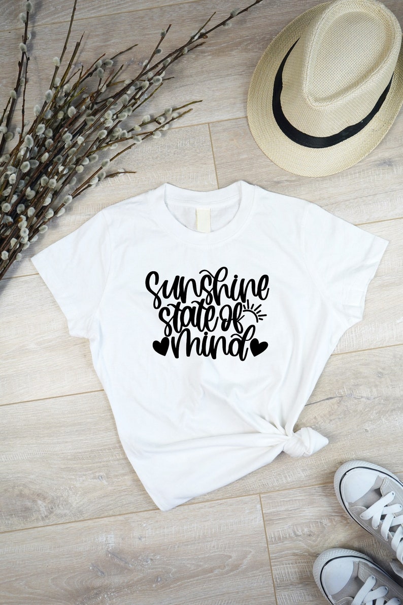 以下が含まれることがあります： 「Sunshine state of mind」という黒い文字が書かれた白いTシャツ。小さな太陽のグラフィックと2つのハートが描かれている。
