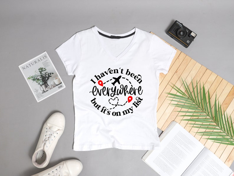May include: White v-neck t-shirt with a black graphic that says "I haven't been everywhere but it's on my list" with a plane and red pins.