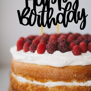 Puede incluir: Un pastel de dos pisos decorado con glaseado blanco y frambuesas frescas. El pastel est&aacute; cubierto con un adorno de pastel negro "Happy Birthday".