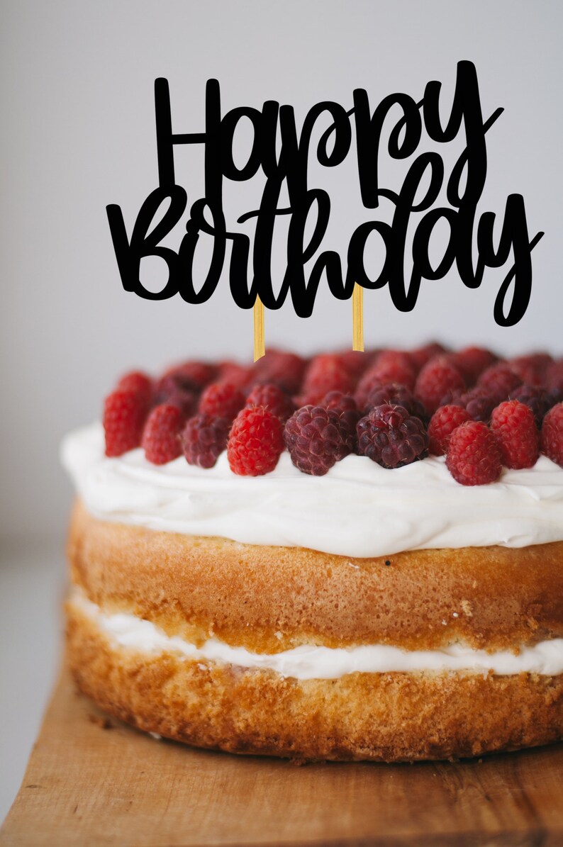 Puede incluir: Un pastel de dos pisos decorado con glaseado blanco y frambuesas frescas. El pastel est&aacute; cubierto con un adorno de pastel negro "Happy Birthday".